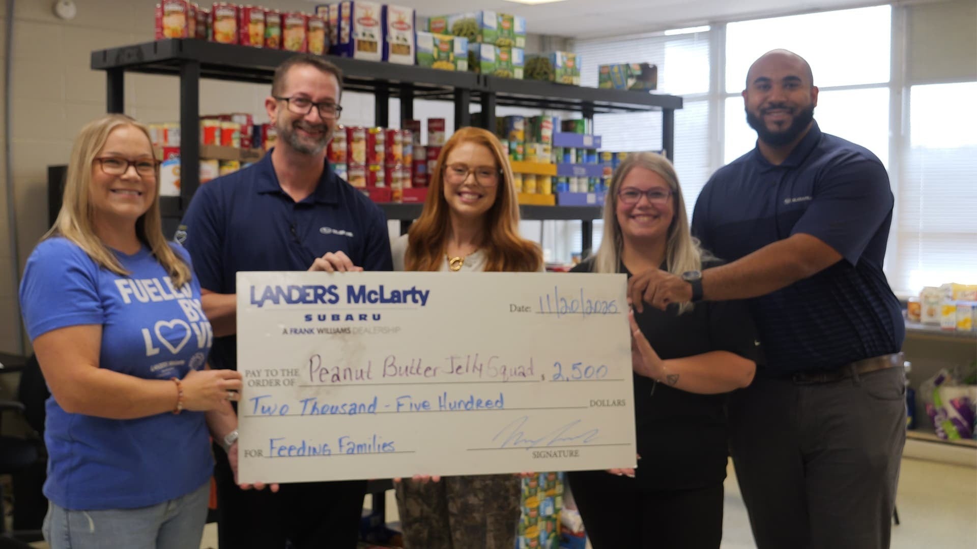 Five smiling people hold a giant check for $2,500 from Landers McLarty Subaru to Peanut Butter Jelly Squad