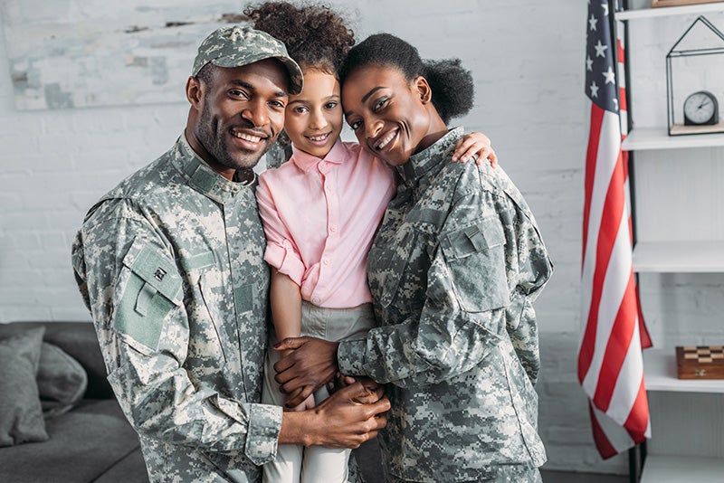Two individuals in military camouflage uniforms standing with a child in a home setting, with a U.S. flag visible in the background